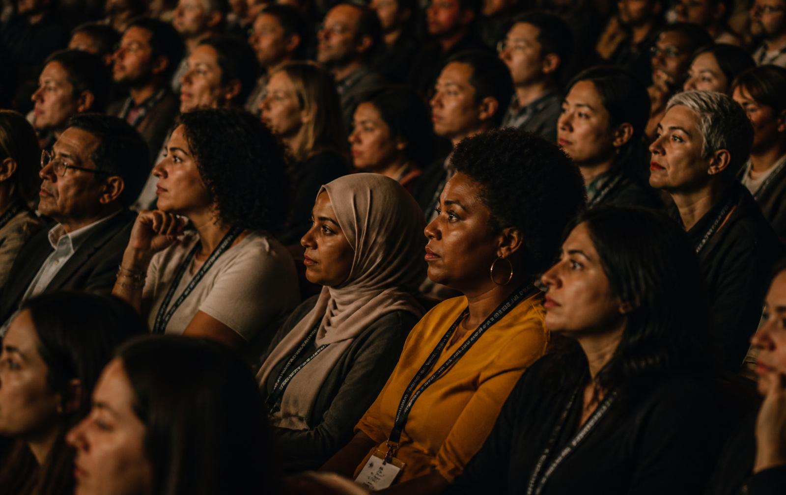 A visibly diverse audience of many ages and backgrounds, lit by warm light, watching intently