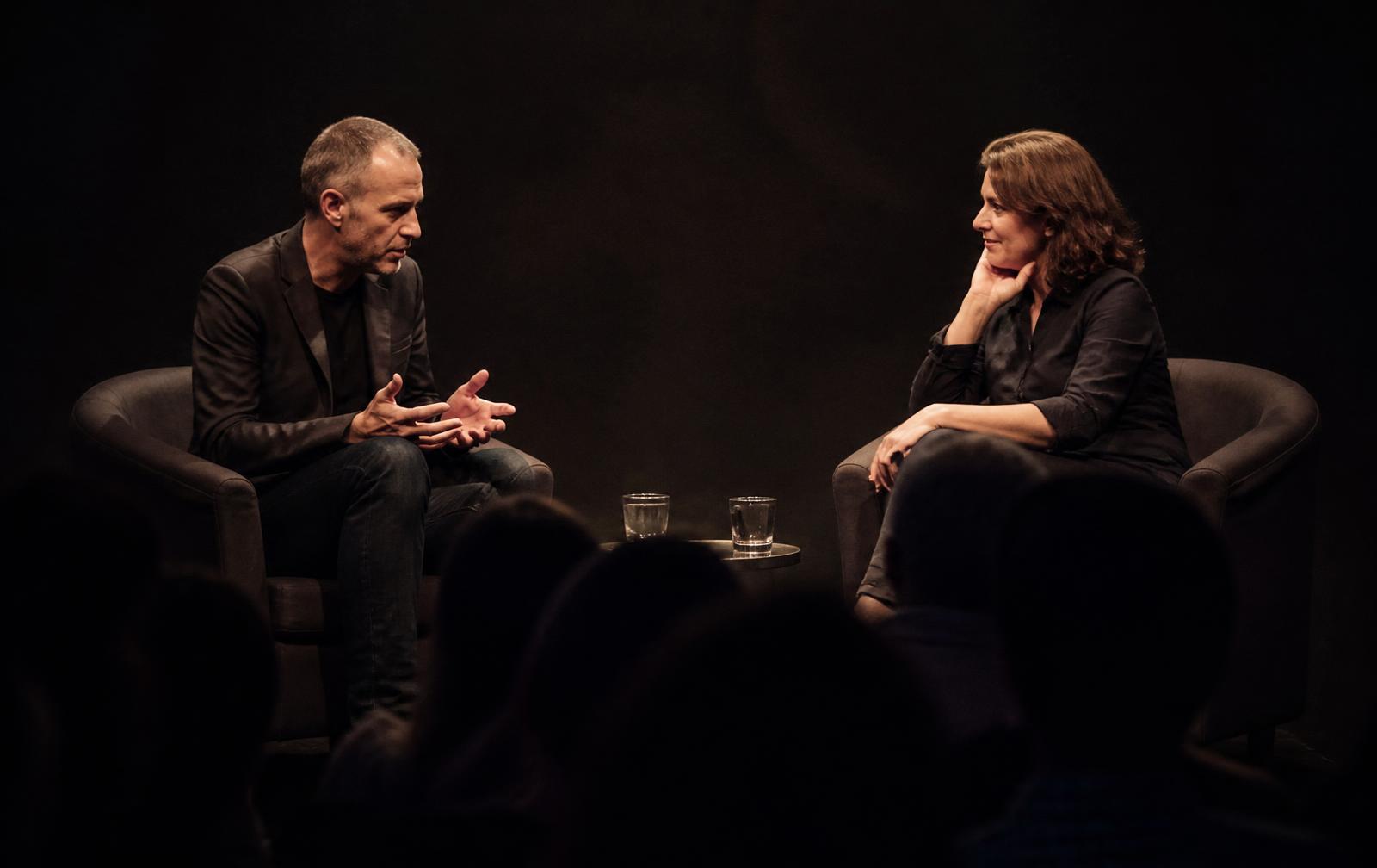 Two people in conversation on an intimate dark stage with silhouetted audience